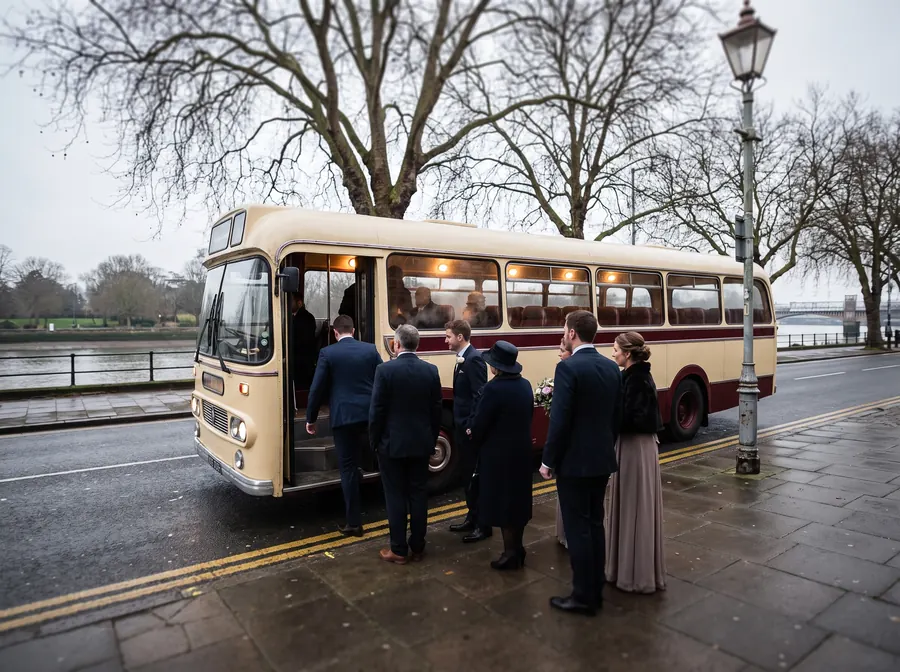A cream and burgundy touring coach parked on the road beside Canbury Gardens on the Thames at Kingston on an overcast winter afternoon, a wedding party in hats and formal dress boarding, bare mature trees and the river behind